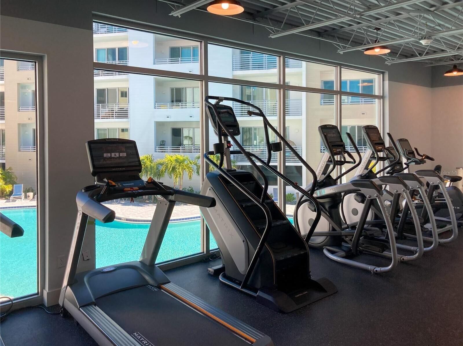 Une salle de sport équipée de tapis de course et de vélos d’exercice, dotée de grandes fenêtres et offrant une vue sur une piscine et des bâtiments.