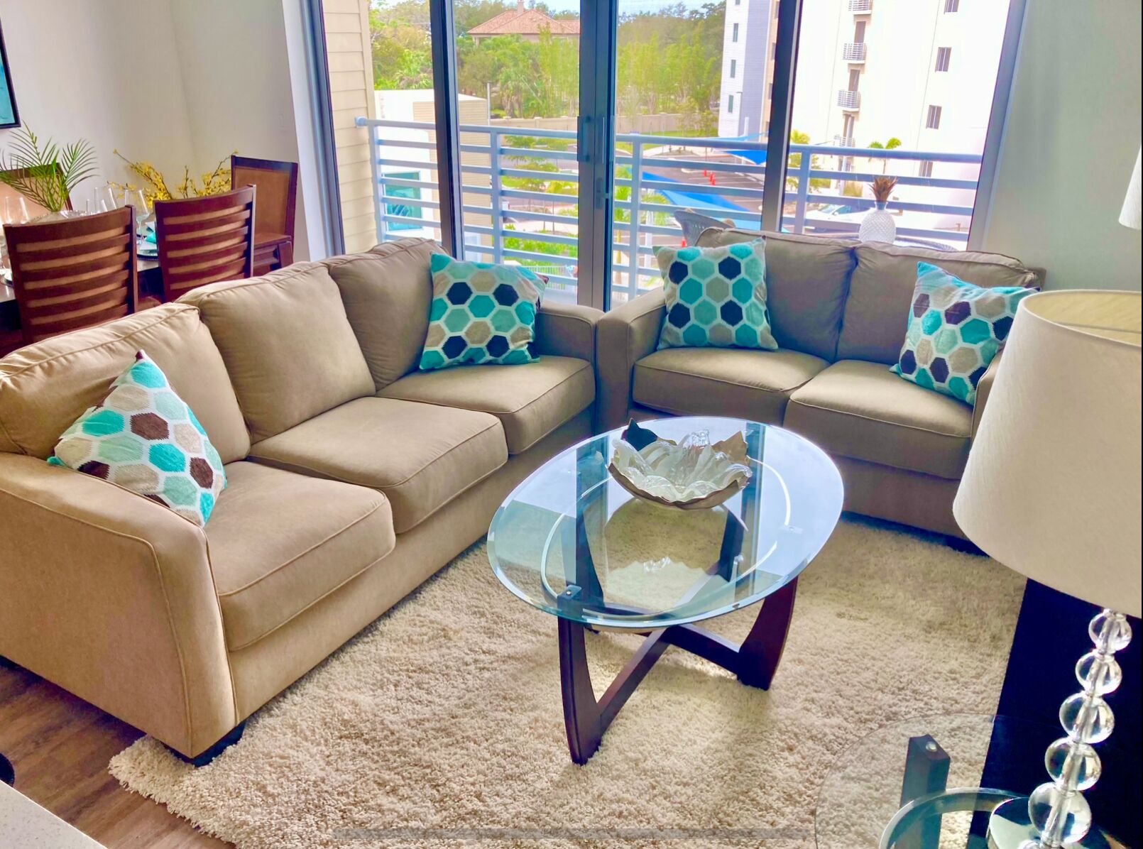 Una sala de estar moderna con un sofá color beige, dos sillones, una mesa de centro de vidrio y vistas a un balcón con edificios y vegetación.