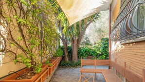 An outdoor patio area with wooden benches and planters, surrounded by greenery and a building with large windows. - Spacious 4 bedroom Apartment near St. Peter's Basilica (Roma)