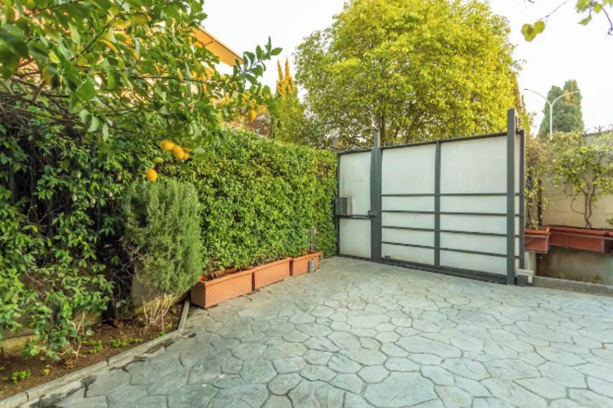 Une allée pavée mène à une entrée fermée par un portail, bordée d’une haie et agrémentée de plantes en pot.