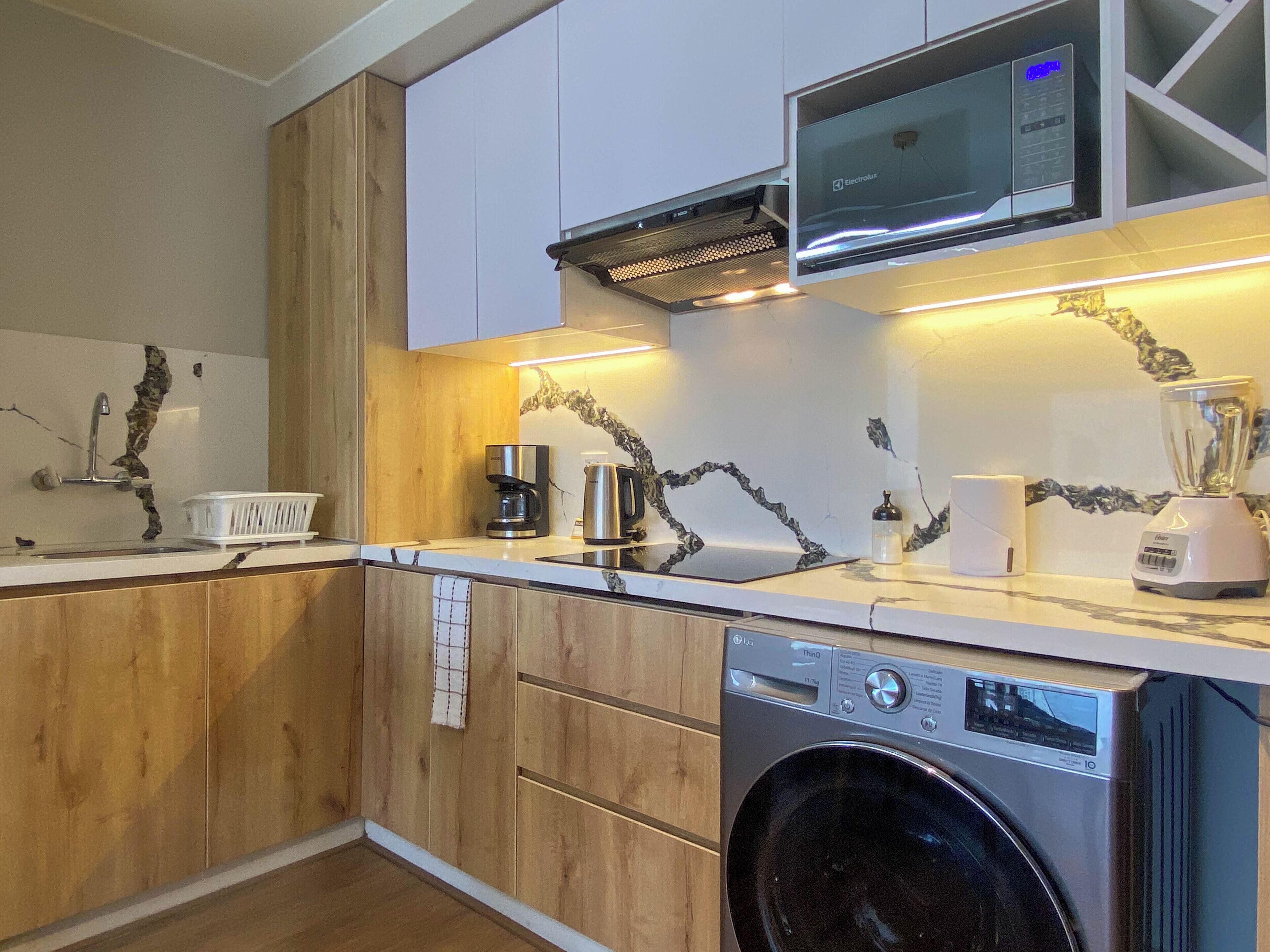 A compact kitchen with wooden cabinets, a built-in microwave, a coffee maker, a blender, and a washing machine.