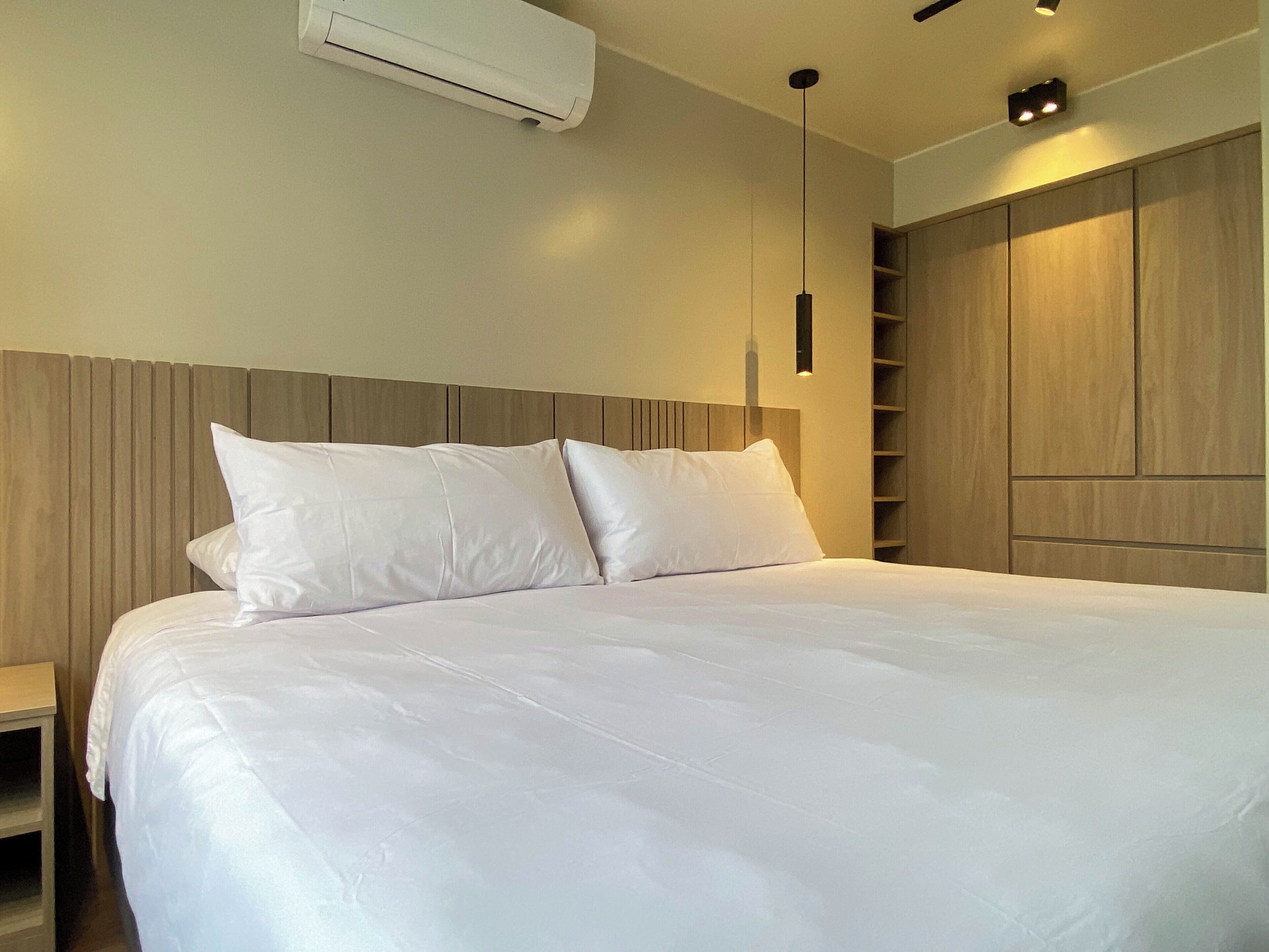 A modern bedroom with a large bed, a wooden headboard, a built-in wardrobe, and a ceiling-mounted air conditioning unit.