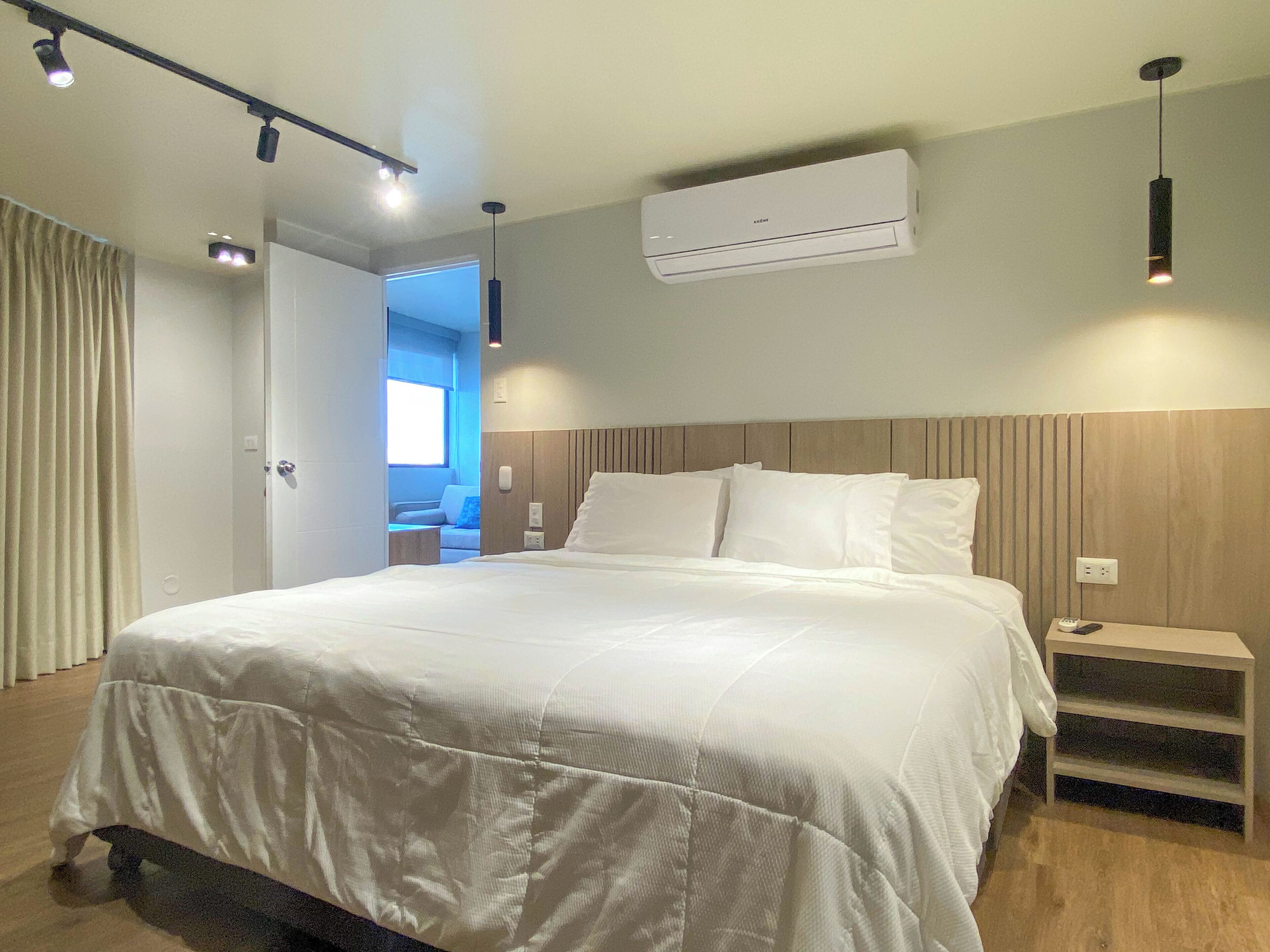 A modern hotel room with a large bed, bedside tables, an air conditioning unit, and a view of the hallway.