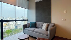 A modern living room with a grey sofa, a round wooden side table, and a brown ottoman. A large window offers a view of a cityscape.