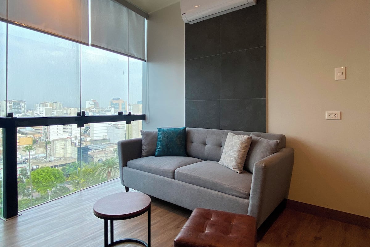 Un salon moderne avec un canapé gris, une petite table d’appoint ronde en bois et un pouf marron. Une grande fenêtre offre une vue sur le paysage urbain.