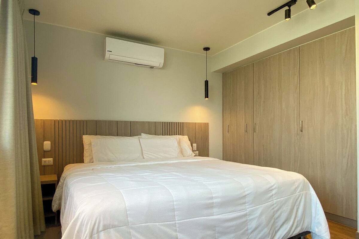 Un lit bien fait dans une chambre d’hôtel, avec une tête de lit en bois, une armoire et un appareil de climatisation fixé au mur.
