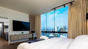 A modern hotel room with a large bed, a flat-screen TV mounted on the wall, a desk with a chair, and a city view through large windows.