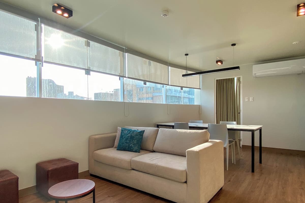 Un salon moderne avec un canapé beige, une petite table ronde et un coin repas comprenant une table et des chaises. De grandes fenêtres laissent entrer la lumière naturelle et offrent une vue sur la ville.