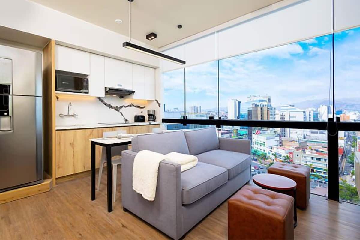 Un salon moderne avec un canapé gris, une petite table et une table basse. On peut admirer une vue sur la ville à travers de grandes fenêtres.