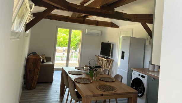 A dining area with a wooden table, chairs, and a TV. There is a kitchen with a washing machine and a refrigerator. - Location Atypique Dans un Airial Landais (Mimizan)