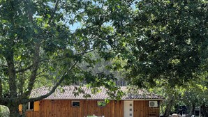 A wooden building with a tiled roof, a door, and windows. There is a green trash bin and outdoor furniture. - Location Atypique Dans un Airial Landais (Mimizan)