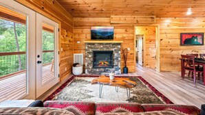 Un interior de cabaña acogedor con chimenea de piedra, paredes de madera, comedor y vistas del bosque a través de amplios ventanales.