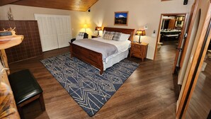 Un dormitorio con techo de madera, una cama con colcha estampada, dos lámparas de noche, una mesita de noche, un banco y vistas a un comedor.