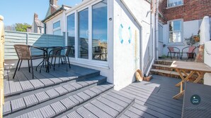 A covered patio with outdoor furniture, a wooden deck, and a brick building in the background. - Harvest Moon - cottage close to harbour (Weymouth)