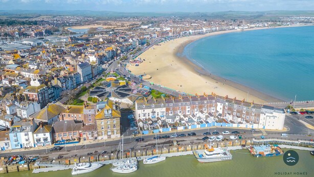 A coastal town with a sandy beach, a marina with boats, and a row of terraced houses along the waterfront. - Harvest Moon - cottage close to harbour (Weymouth)