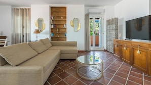 Una sala de estar con un sofá seccional color beige, una mesa redonda, una estantería, un televisor y un ventanal corredero que da a un balcón.