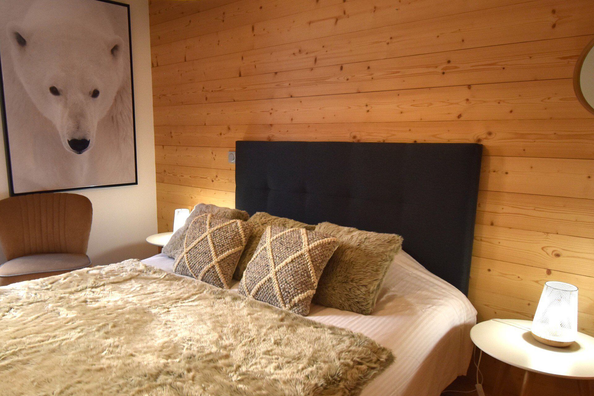 A hotel room with a wooden wall, a bed with a dark headboard, two patterned pillows, a fluffy blanket, a chair, a small table with a lamp, and a framed picture on the wall.