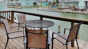 A wooden deck with a round table and four chairs, overlooking a calm water body with buildings in the background. - Changes in Attitudes by Miss Kitty’s (Rockport)