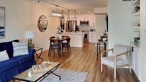 A modern living room with a blue sofa, a coffee table, and a dining area with a chandelier. - Changes in Attitudes by Miss Kitty’s (Rockport)