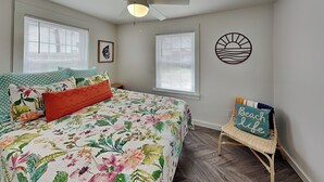 A bedroom with a floral bedspread, a ceiling fan, two windows with blinds, a wall art of a sun and waves, and a beach-themed pillow on a chair. - Coral Cottage by Miss Kitty’s (Rockport)
