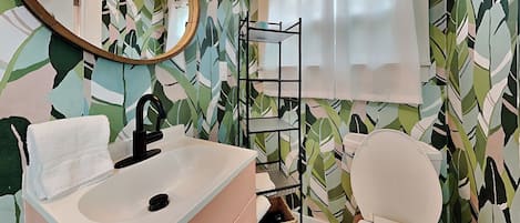 Un baño con un lavamanos blanco, grifo negro y un inodoro blanco. Hay un espejo sobre el lavamanos y un estante con una cesta debajo. Las paredes están decoradas con un diseño de hojas.