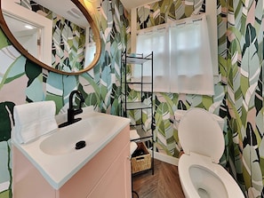 A bathroom with a white sink, black faucet, and a white toilet. There is a mirror above the sink and a shelf with a basket below it. The walls are decorated with a leafy pattern.