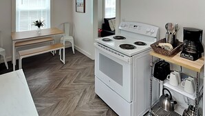 A compact kitchen with a white stove, a coffee maker, and a small dining area with a bench and chairs. - Coral Cottage by Miss Kitty’s (Rockport)