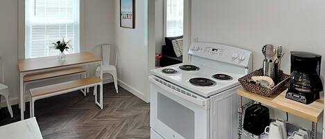 Una cocina compacta con estufa blanca, cafetera y un pequeño comedor con banco y sillas.