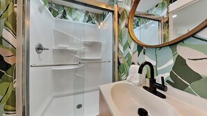 A modern bathroom with a glass-enclosed shower, a white sink with a black faucet, and a mirror with a wooden frame. - Coral Cottage by Miss Kitty’s (Rockport)