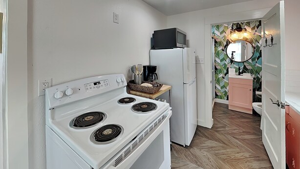 A compact kitchen with a white stove, a refrigerator, and a microwave. There's a small countertop with a coffee maker and utensils. The kitchen has a herringbone-patterned backsplash and a wooden floor. - Coral Cottage by Miss Kitty’s (Rockport)