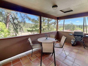 A covered patio with a round table and chairs, a grill, and a view of the surrounding landscape. - The Bungalow by Ruidoso Vacation Rentals (Ruidoso)