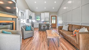 A modern living room with a sofa, armchairs, and a coffee table. There is a kitchen area with white cabinets and a dining table.