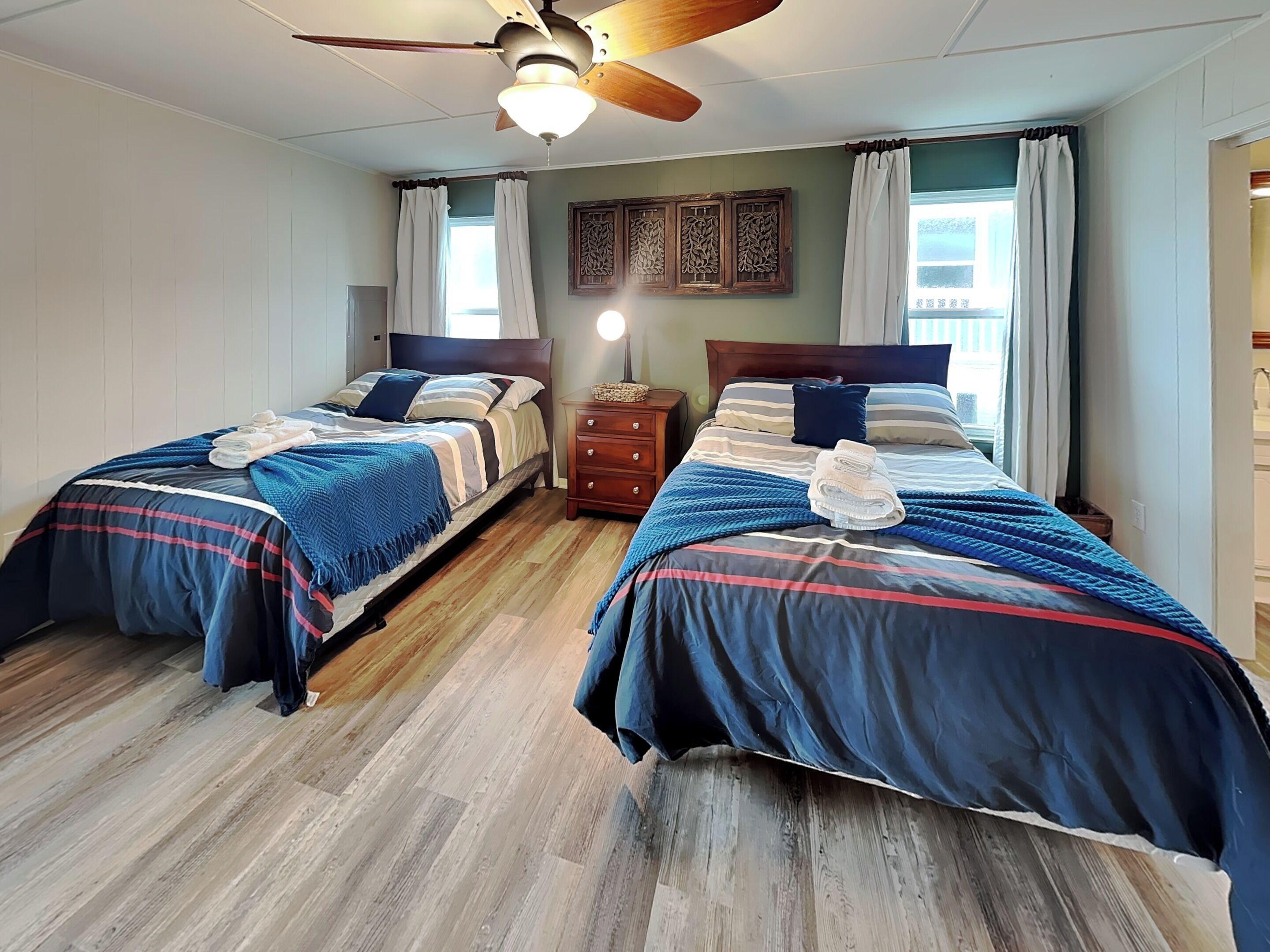 A room with two beds, wooden flooring, a ceiling fan, and a nightstand with a lamp.