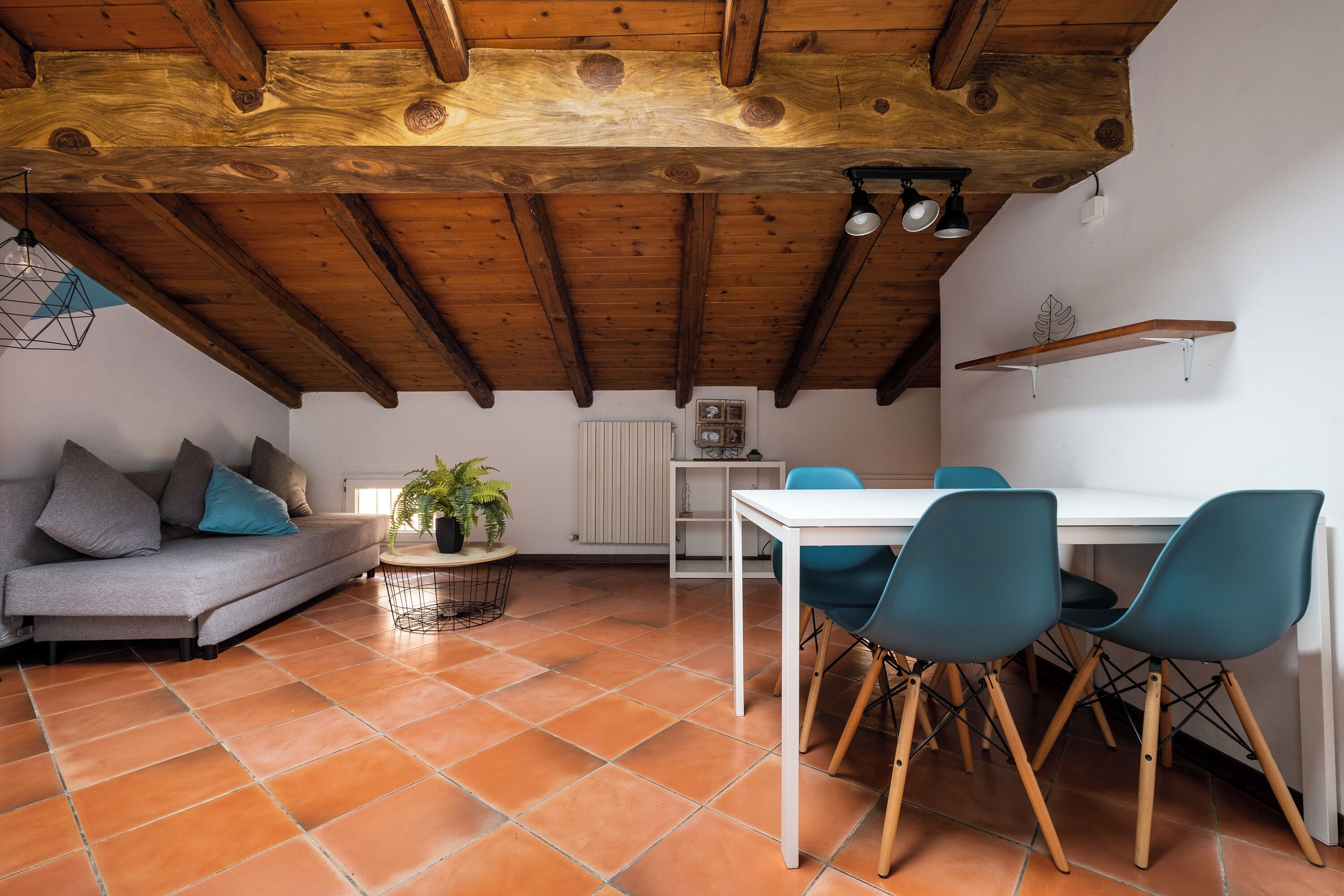 A cozy living area with a wooden ceiling, a sofa with cushions, a dining table with chairs, and a potted plant.