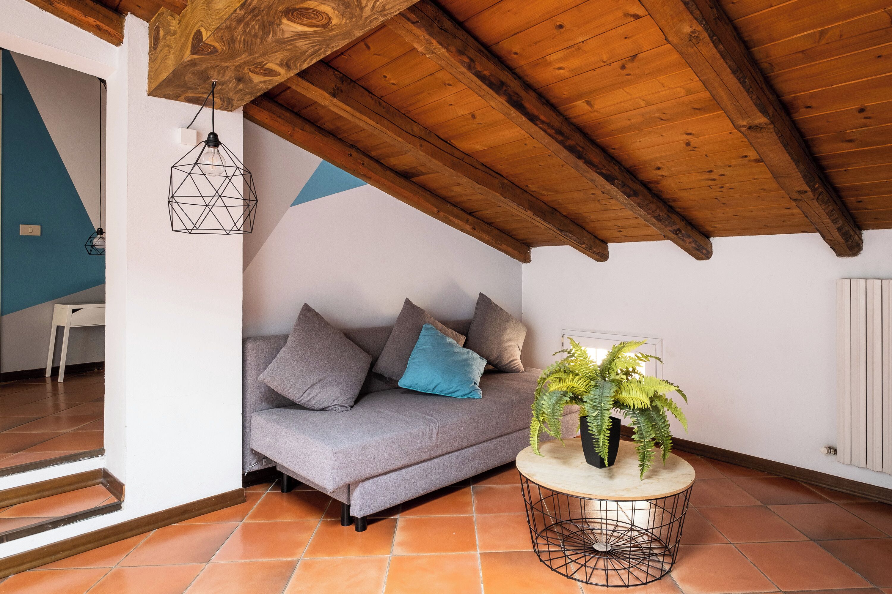 Een gezellige kamer met een houten plafond, een grijze bank met kussens, een rond bijzettafeltje met een plant en een geometrische hanglamp.
