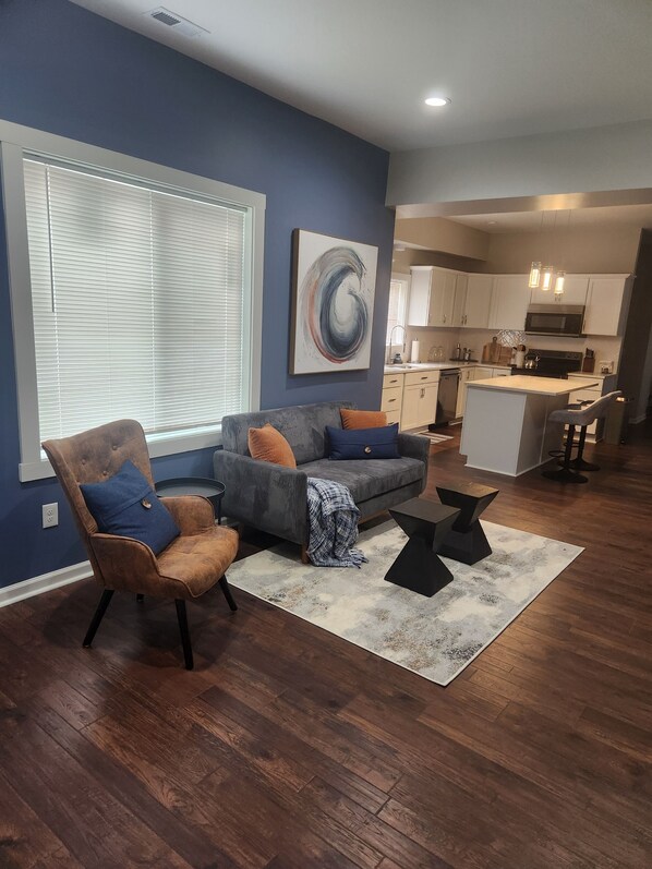 A modern living room with a sofa, armchair, and coffee table. A kitchen is visible in the background with white cabinets and a microwave. - Cozy 3-bedroom house near Lucas Oil Stadium in downtown Indianapolis.  (Indianapolis)