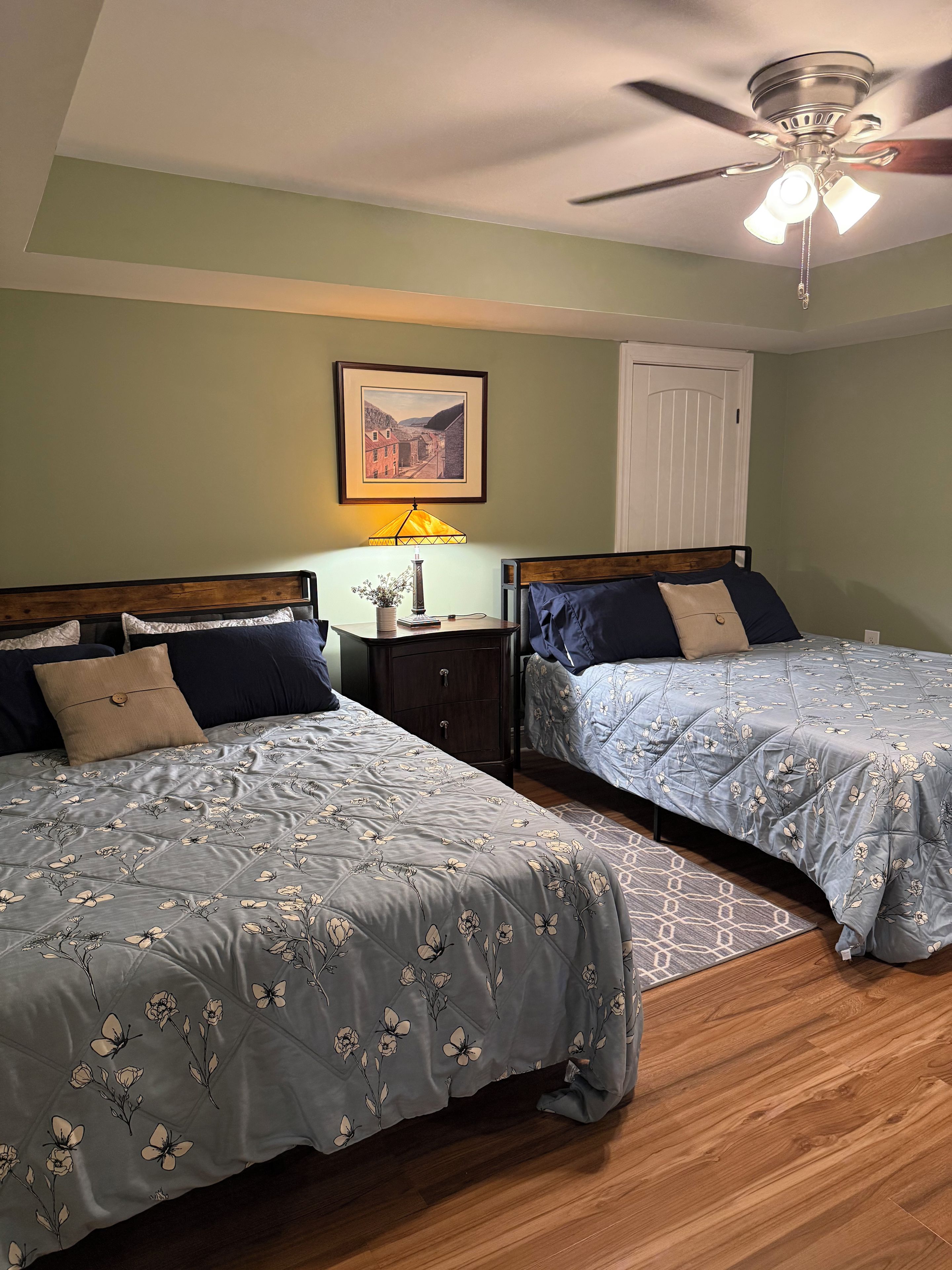 A bedroom with two beds, a ceiling fan, a nightstand with a lamp, and a framed picture on the wall.
