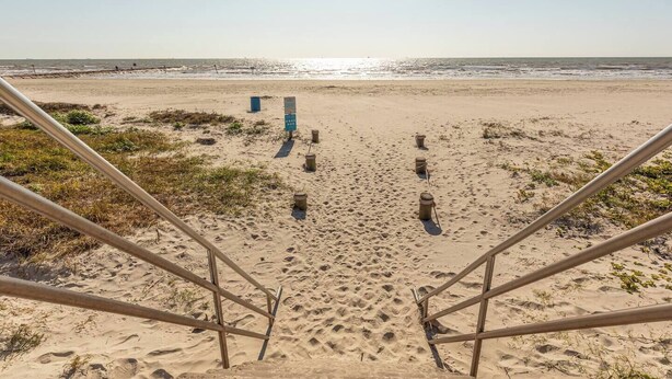 A sandy beach with a metal staircase leading down to the water, a blue lifeguard stand, and several empty buckets lined up in the sand.