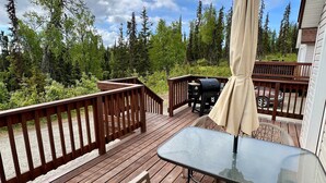 A wooden deck with a glass table and chairs, a barbecue grill, and a covered umbrella. - Peaceful Acres Cabin 2 - Eagle's Nest  (Soldotna)