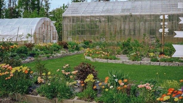 A greenhouse with a transparent roof, surrounded by a variety of flowering plants and a well-maintained garden. - Peaceful Acres Cabin 3 - Cozy Cottage  (Soldotna)