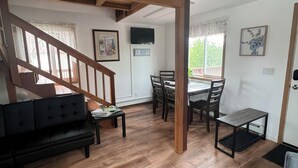 A spacious living area with a wooden ceiling, a black sofa, a dining table with chairs, and a staircase leading to another level. - Peaceful Acres Cabin 3 - Cozy Cottage  (Soldotna)