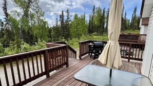 A wooden deck with a glass table and chairs, a beige umbrella, and a view of trees and sky. - Peaceful Acres Cabin 3 - Cozy Cottage  (Soldotna)