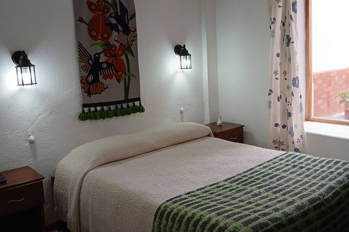 Un dormitorio con cama, dos mesitas de noche, un tapiz colgado en la pared, dos lámparas de pared, un ventilador de techo y una ventana con cortinas.