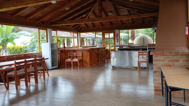 Un espacio interior amplio con techo alto, vigas de madera, una zona de cocina con horno de ladrillo y grandes ventanales que ofrecen vistas al verdor.