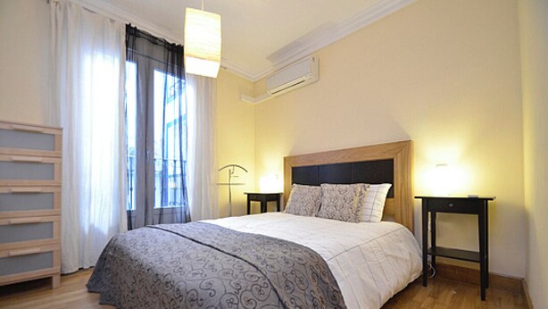 A bedroom with a wooden bed, a bedside table, a lamp, and a window with curtains.