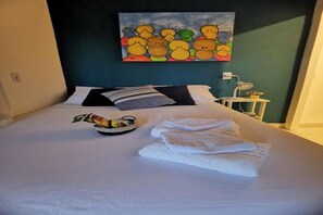 A neatly made bed with white linens, a basket of fruits, and a painting on the wall.