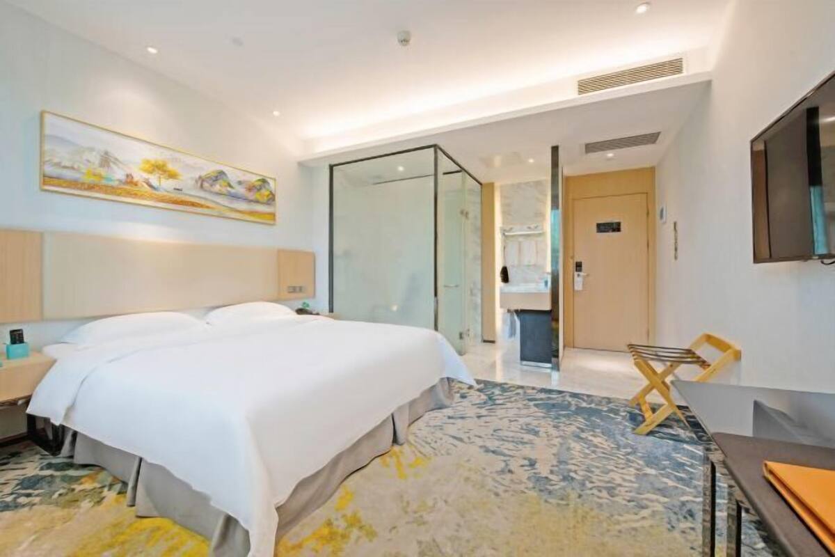 A modern hotel room with a large bed, a glass partition, a television, and a painting on the wall.