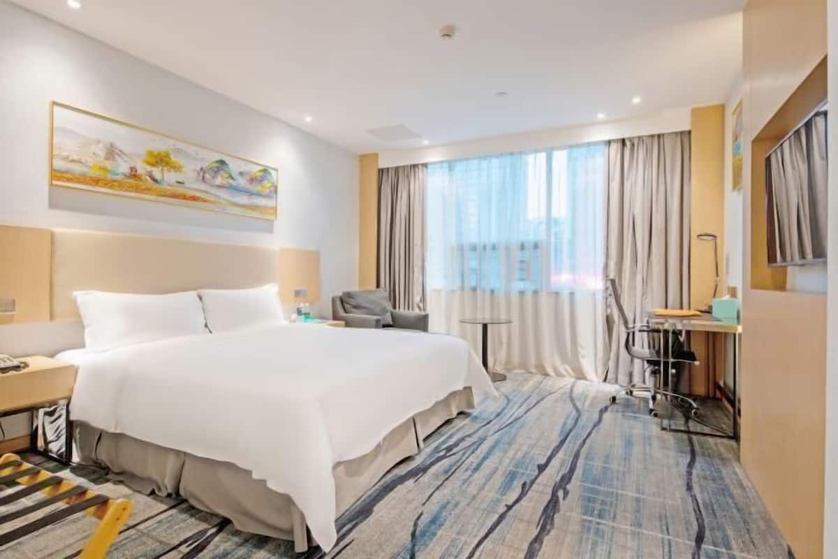 A hotel room with a large bed, a desk, a chair, a small table, and a window with curtains.