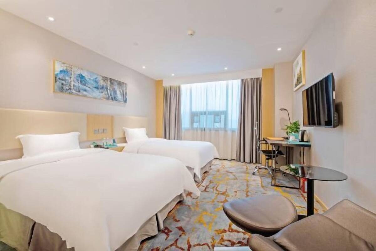A hotel room with two beds, a desk, a chair, a TV, and a large window with curtains.