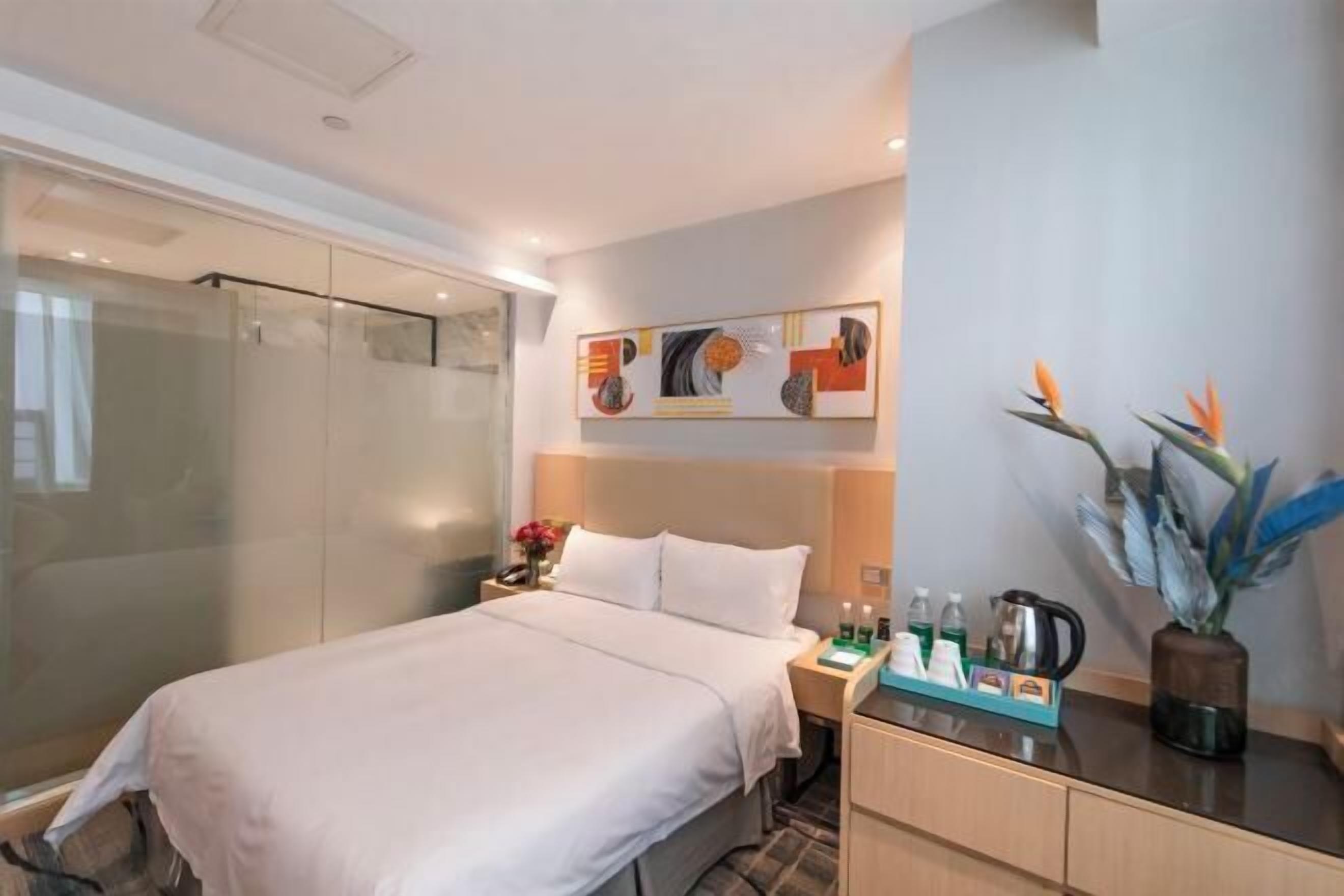 Una habitación de hotel con cama, mesita de noche, una obra de arte en la pared, un jarrón con flores y un box de ducha de vidrio.